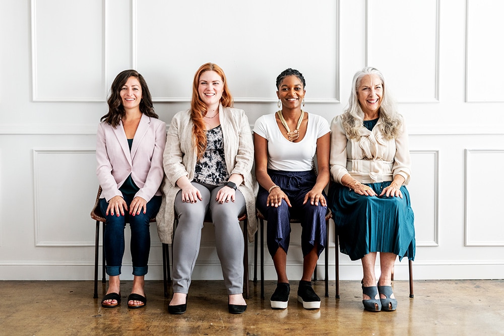 Menopause care Forest Hill, A group of five women sitting on chairs against a white wall, appearing relaxed and engaged with one another